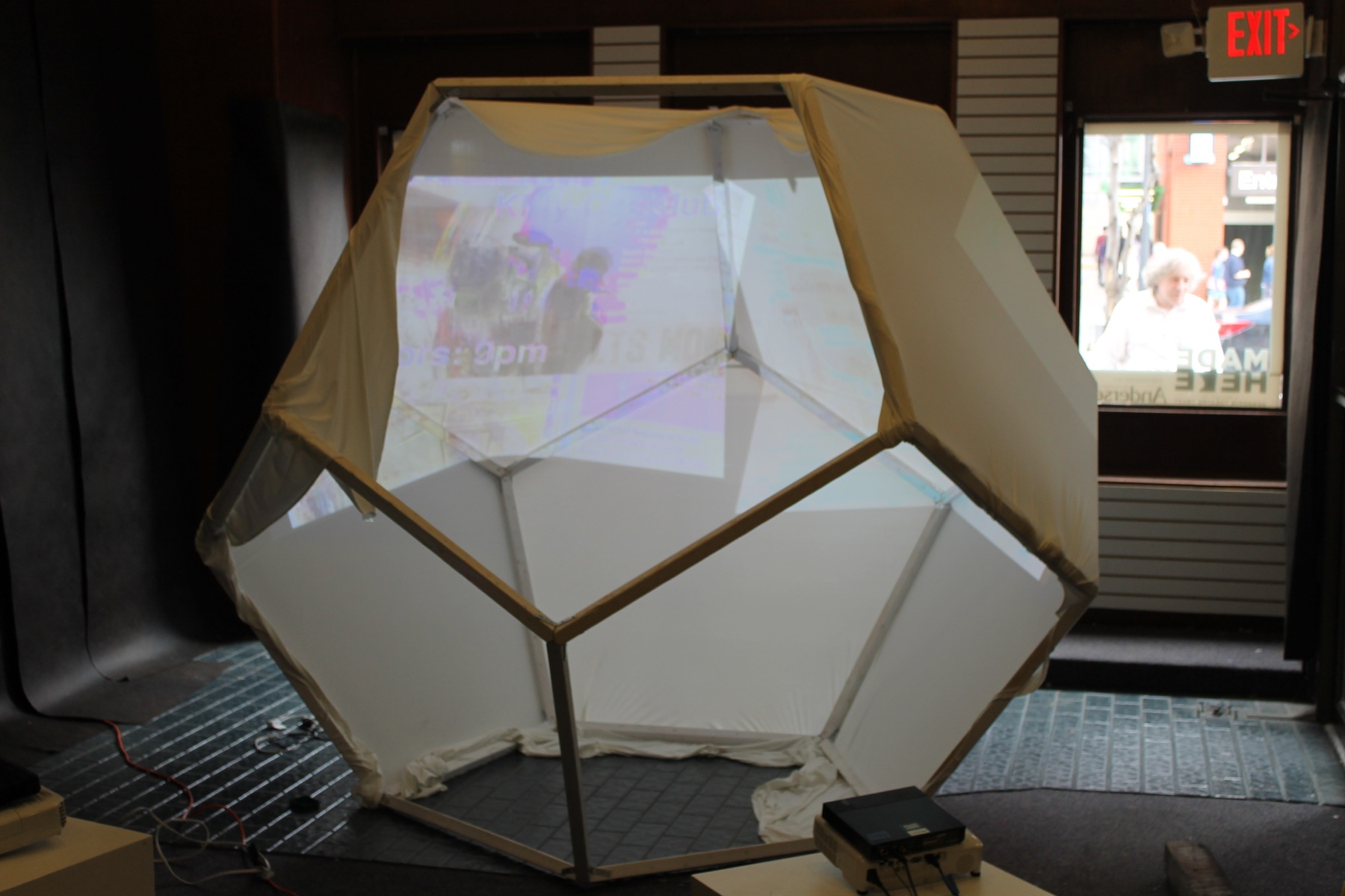 Faceted white projection structure installed in a storefront, with a pale moving image cast across its folded surfaces.