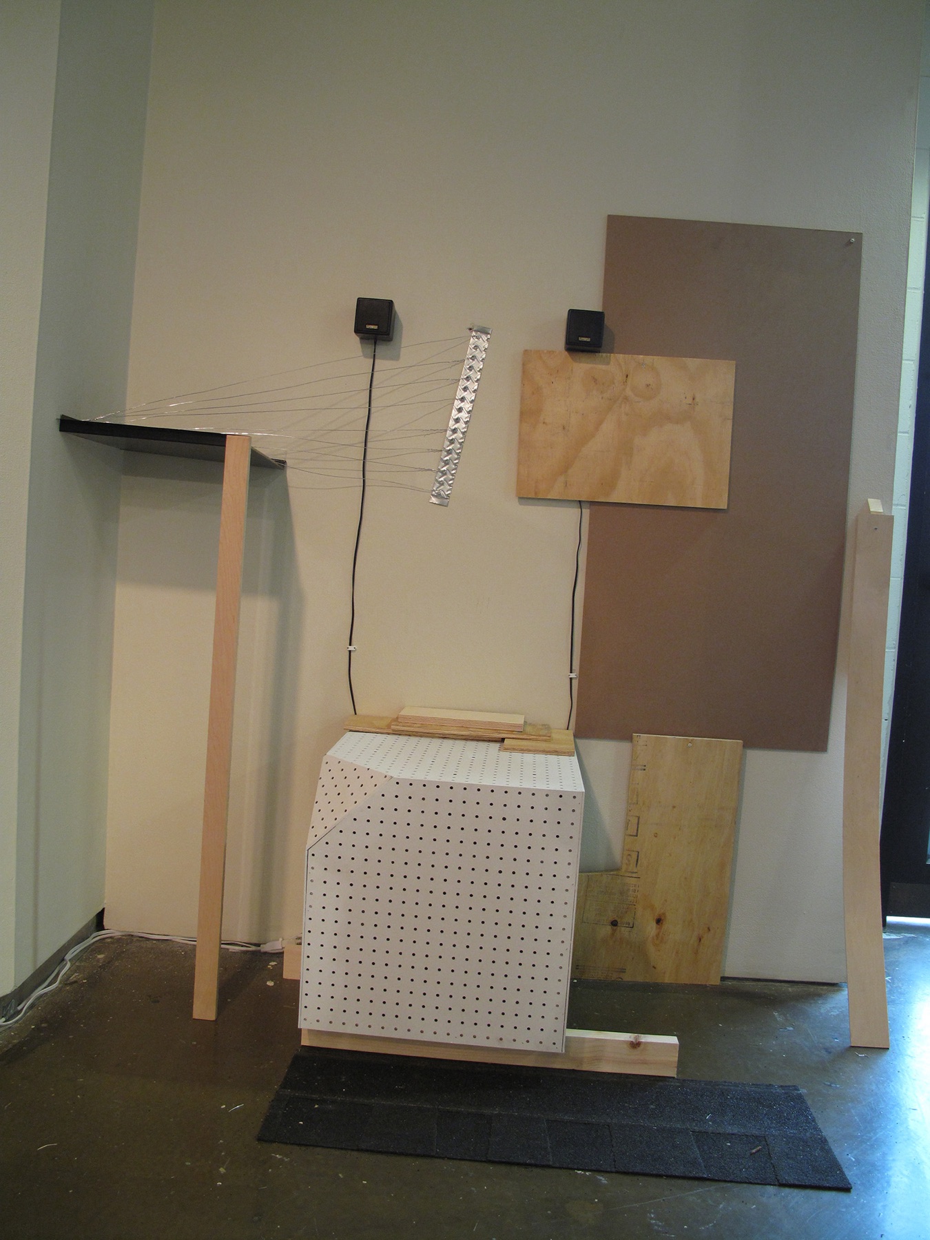 Plywood, perforated white panels, wood strips, and two small black components assembled against a gallery wall.
