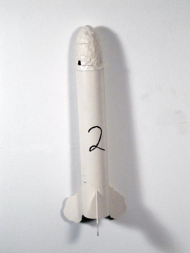 White model rocket marked with a handwritten 2, with a taped nose cone and small fins, mounted against a white wall.