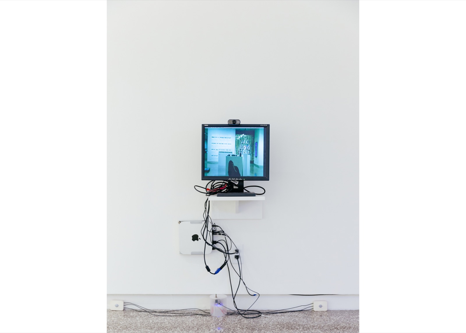 Wall-mounted monitor and webcam above a compact computer, control enclosure, visible cabling, and two small sensor boxes placed along the floor.